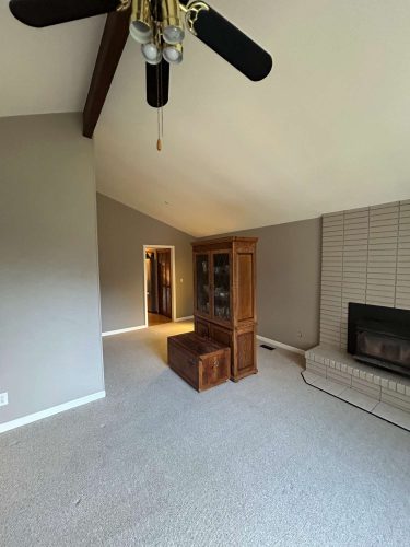 A cozy living room painted by Pearl Painters in Oneal, Portland, features beige carpet, gray walls, a wooden cabinet and chest, brick fireplace, ceiling fan with lights, and a vaulted ceiling with exposed beam.