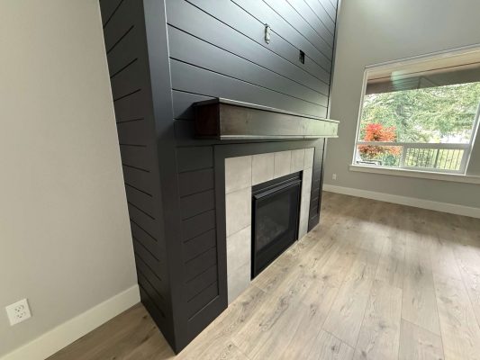 Modern interior living room in Portland with a black shiplap fireplace, wood mantel, tiled surround, light wood floor, and large window. Painted by Pearl Painters.