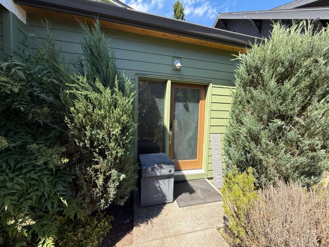 A green building in Portland, with a glass door, tall bushes, a trash bin, and a delivery package at the entrance is shown under sunny May 2025 skies. Pearl Painters can provide professional painter and painting services for such exteriors.