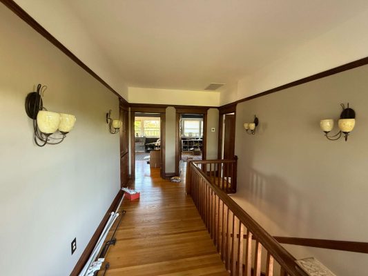 This second-floor interior hallway in a Dennis home features hardwood floors, a wooden railing, beige walls, and wall-mounted lights. Items are on the floor as of May 2025. No painting or painter work by Pearl Painters of Portland is noted here.