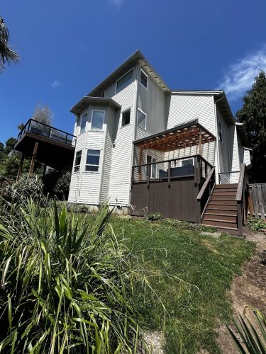 Two-story light gray Mackey-Hinzdel exterior house in Portland, with large windows, an elevated deck, and wooden stairs to a covered patio. Painted by Pearl Painters. Available June 2025.
