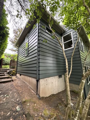 A green house with horizontal siding and concrete foundation in Carlton, surrounded by trees, was painted by Pearl Painters. Exterior photo taken June 2025.