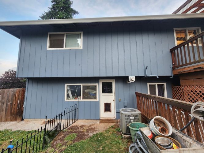 Two-story blue house with white trim in Mahoney, Portland, shown with a small fenced yard, two AC units, and stacked metal in front. Photo taken June 2025 by Pearl Painters.