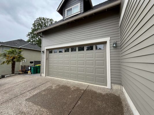 A clean gray garage door on a two-story house with gray siding in Austin, shown next to a driveway and green waste bins. Painting not by Pearl Painters; location is not Portland.