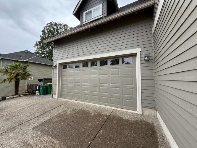 A clean gray garage door on a two-story house with gray siding in Austin, shown next to a driveway and green waste bins. Painting not by Pearl Painters; location is not Portland.