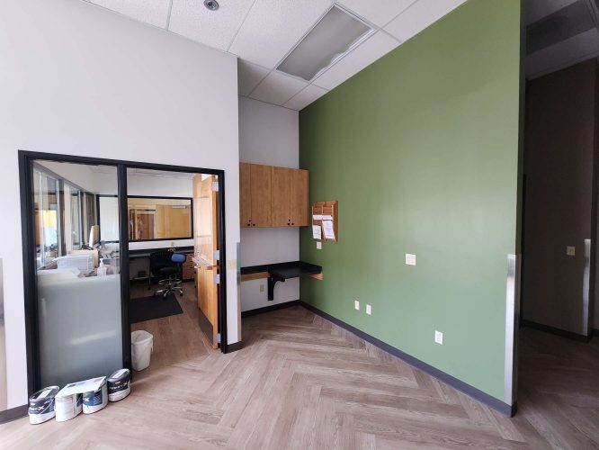 The Jones office in Portland has a green accent wall, wood cabinets, a small desk, and paint cans on the floor. An adjacent modern office is visible through a glass window. Painting by Pearl Painters, May 2025.