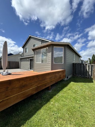 A two-story gray house with white trim—recently painted by Pearl Painters—features a stylish exterior and sits behind a raised wooden deck in a fenced backyard in Portland.