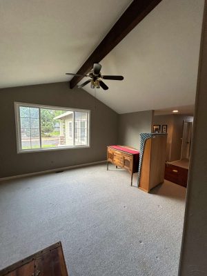 This carpeted room in Portland features gray walls, a large window, ceiling fan, wooden Oneal furniture, and a sloped ceiling with exposed beams. Pearl Painters' painting work is evident throughout the space.