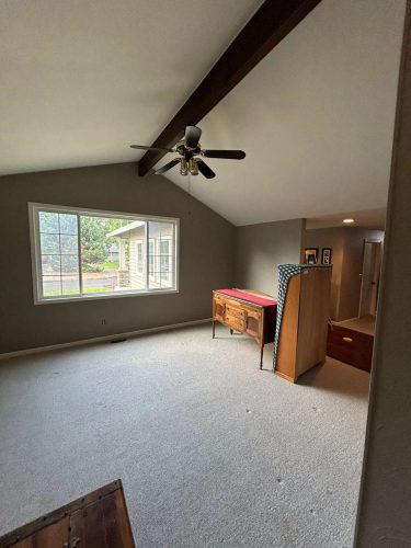 This carpeted room in Portland features gray walls, a large window, ceiling fan, wooden Oneal furniture, and a sloped ceiling with exposed beams. Pearl Painters' painting work is evident throughout the space.