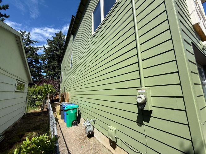 The green exterior side wall of the Portland house, painted by Pearl Painters, shows utility meters, recycling and trash bins, and a narrow fenced side yard (photo: May 2025).
