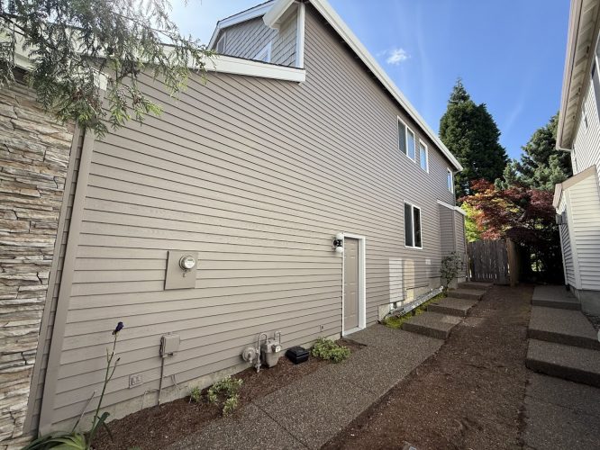 A Pearl Painters painter photographed a beige house with horizontal siding, utility meters, and a garden bed along a walkway in Hendricks, not Portland, May 2025.