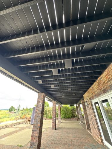A painting by Pearl Painters depicts a covered patio with dark wooden beams and brick columns at Anderson Extension, Portland, June 2025, overlooking an open outdoor area.