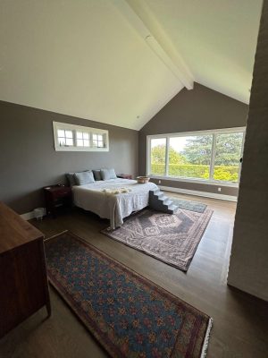 A modern bedroom in Portland, designed by Duffy, features a large bed, area rugs, and wooden furniture. Floor-to-ceiling windows showcase greenery. Pearl Painters provided painting services for this space.