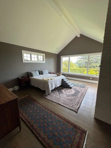 A modern bedroom in Portland, designed by Duffy, features a large bed, area rugs, and wooden furniture. Floor-to-ceiling windows showcase greenery. Pearl Painters provided painting services for this space.
