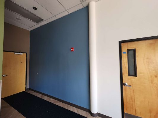 Portland painter Pearl Painters painted a plain hallway interior with a blue accent wall, red alarm, closed wooden door labeled "Jones office," black mat, and high white ceiling with recessed lighting.