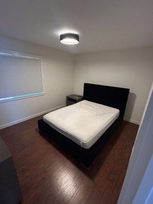 A neatly made bed with a black frame and headboard sits in a bedroom with wood flooring, a nightstand, and a covered window. Painted by Pearl Painters, Portland.