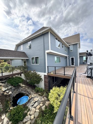 This modern two-story Anderson home in Portland features blue siding, white trim, a wooden deck walkway with metal railings, and landscaped rocks. Pearl Painters completed the painting. Ready for move-in June 2025.