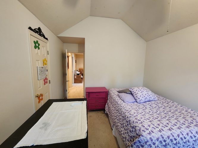 A small bedroom at Haase Ext. features a single bed with purple floral bedding, a pink nightstand, a dresser with padding, and a decorated door to the hallway. Ideal for relaxing or studying. For painting needs in Portland, contact Pearl Painters.