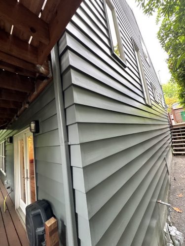 Close-up of a grey house with horizontal siding, window, door, and part of a wooden deck in Carlton. Painting by Pearl Painters, captured June 2025.