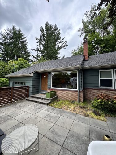 This single-story house in Carlton features blue-green siding, a wood door, brick chimney, and a fenced patio with outdoor furniture. The painting shows trees and a cloudy June 2025 sky. Painted by Pearl Painters, Portland painter.