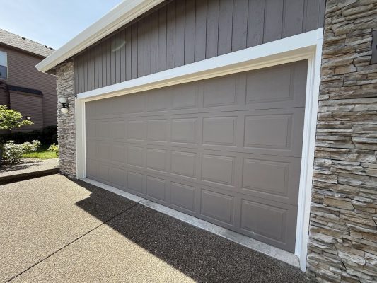 Closed gray Hendricks garage door on a modern house with stone accents and brown siding in bright sunlight, Portland. Photo by Pearl Painters, May 2025.