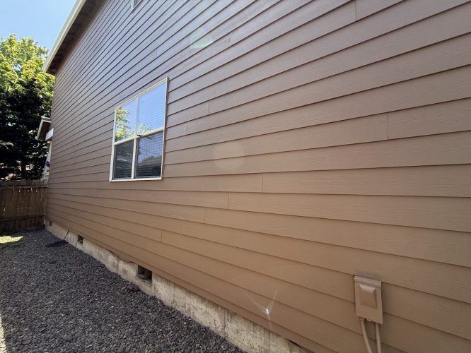 Side view of a tan house with horizontal siding, one window, and a utility box near the base. Gravel surrounds the exterior. Trees and a wooden fence are visible. Painted by Pearl Painters in Emory, June 2025.