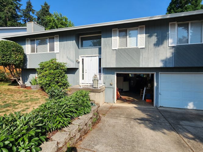 A gray split-level Mahoney house with white trim, white front door, single-car garage, and concrete driveway with plants and a small lawn. Located in Portland. Available for exterior painting by Pearl Painters in June 2025.