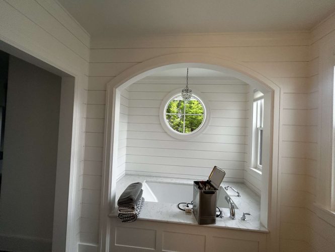 A white shiplap-walled bathroom in Portland, styled by Duffy Interior for May 2025, features a marble-topped bathtub, round window with greenery view, small chandelier, and folded towels on the tub edge. Ideal painting inspiration for Portland painters like Pearl Painters.
