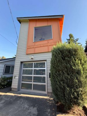 A two-story Arain house in Portland features white siding, an orange accent, large window, glass garage door, and a tall bush in front. Painted by Pearl Painters, this modern painting will be showcased in May 2025.
