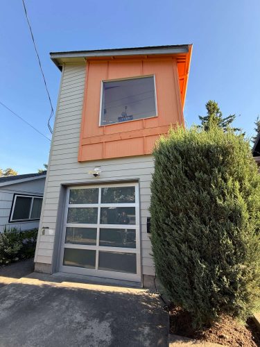 A two-story Arain house in Portland features white siding, an orange accent, large window, glass garage door, and a tall bush in front. Painted by Pearl Painters, this modern painting will be showcased in May 2025.