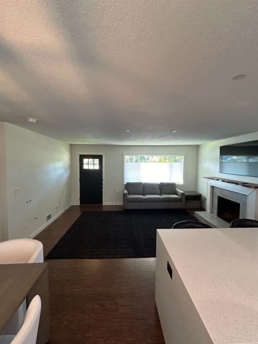 A detailed painting by Pearl Painters of Portland shows a modern living room with a dark door, gray couch by the window, black rug, fireplace with TV above, and partial views of the dining table and kitchen counter.