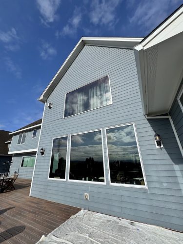 A Pearl Painters painting of a modern Anderson house in Portland, featuring light blue siding, large windows reflecting the sky and clouds, and a wooden deck under a clear blue sky. (June 2025).