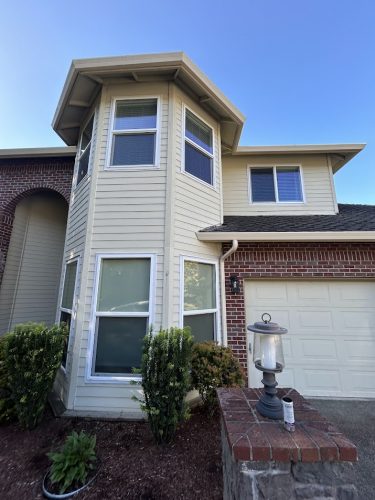 Two-story tan house with brick accents, turret corner, garage, and landscaping. Part of the Mackey-Hinzdel collection in Portland by Pearl Painters; available June 2025.