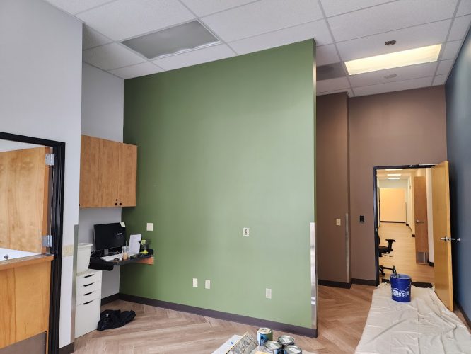 Office in Portland with green accent wall, wooden cabinets, and small desk area. Painting supplies and covered floor indicate ongoing painting by painter Jones as of June 2025.