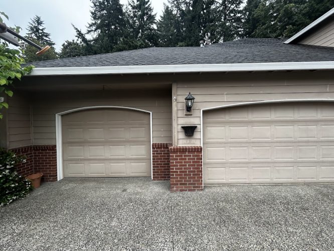 Two beige garage doors with brick accents on a Haase residential house in Portland, painted by Pearl Painters in June 2025. Features include a wall-mounted lantern and a potted plant.