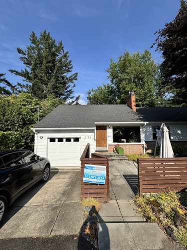A single-story house in Carlton with a gray roof, white garage, parked black car, and a real estate sign on the front wooden fence. Mature trees surround the property. Photo by Pearl Painters, June 2025. Carlton is west of Portland.