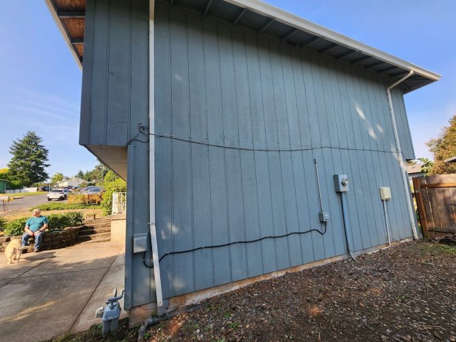 Blue house with vertical siding and visible utility meters in Portland, photographed by Mahoney, June 2025. A man and dog sit on a bench nearby. Pearl Painters offers painter and painting services in the area.