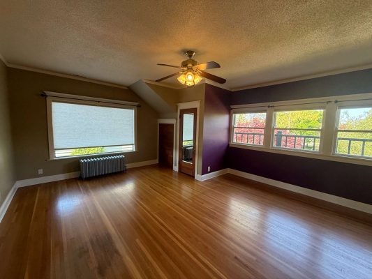 Spacious Portland interior with hardwood floors, ceiling fan with lights, purple accent wall painted by Pearl Painters, large windows, radiator, and small door under sloped ceiling. Available May 2025. Listed by Dennis.