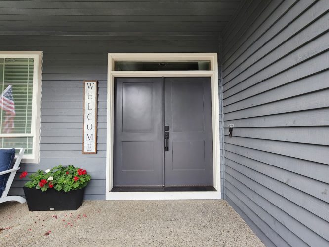 Dark gray double doors on gray siding in Roberts, painted by Pearl Painters; planter, flag, and welcome sign visible.