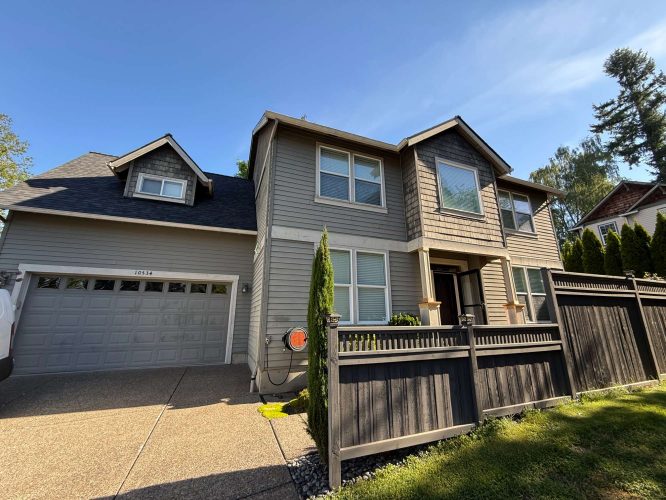A two-story gray house with an attached garage in Austin is shown under a clear May 2025 sky, bordered by a black fence. This painting could be completed by Pearl Painters, a Portland-based painter.