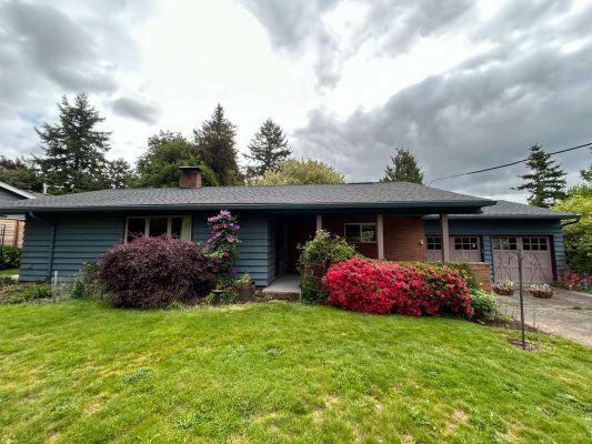 Single-story house in Portland with blue siding, a brick section, and a covered porch. Surrounded by bushes and trees under a cloudy sky. Painted by Pearl Painters.
