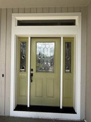 A green front door with decorative glass panels and sidelights, framed by beige siding and white trim, is a painting idea for your Hendricks home. For professional painters in Portland, contact Pearl Painters.