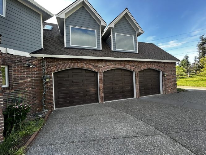 A painter from Pearl Painters painted the brown doors of a three-car Anderson garage attached to a brick and gray-sided house in Portland. The painting was done under a clear blue June 2025 sky.