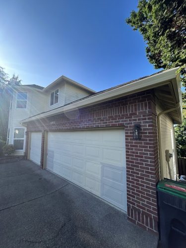 Two-car garage extension attached to a two-story Mackey-Hinzdel home with red brick and beige siding, located in Portland. Driveway in front, with trees and blue sky visible. Painting by Pearl Painters. Available June 2025.