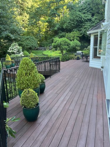 A painting by Herrle depicts a wooden deck with potted topiary shrubs, black metal railings, and outdoor furniture overlooking a backyard garden. This artwork is featured by Pearl Painters, a Portland painter, as an example of outdoor retreat design for June 2025.