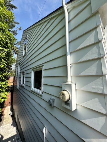 Side view of a Carlton house near Portland with light-colored siding, two small windows, electric meter, and conduit pipes. Photo taken June 2025 by Pearl Painters, local painters specializing in house painting.