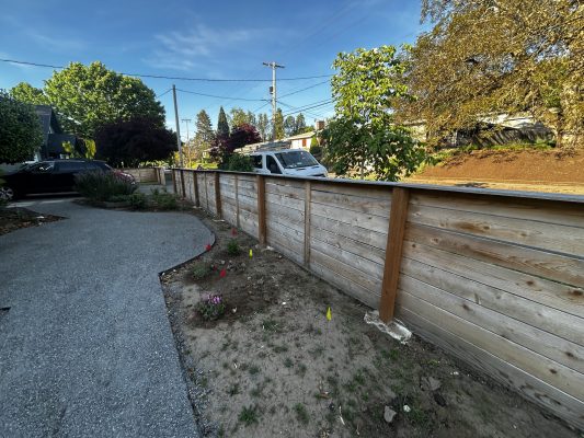 In May 2025 in Trujillo, a painter's white van is parked on the street near an exterior wooden fence by a walkway and flower bed.