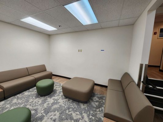 A small waiting area in the Jones office, Portland, features two brown sofas, two green stools, a brown ottoman, a patterned rug, and white walls under fluorescent lights. No people or Pearl Painters are present; no painting is underway.