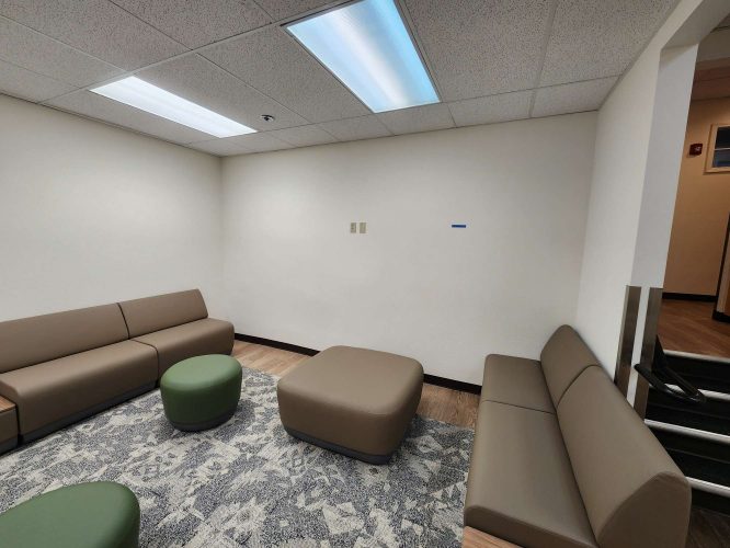 A small waiting area in the Jones office, Portland, features two brown sofas, two green stools, a brown ottoman, a patterned rug, and white walls under fluorescent lights. No people or Pearl Painters are present; no painting is underway.