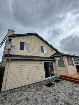 A painter from Pearl Painters in Portland painted this two-story beige house with a satellite dish, sliding glass door, small deck, and paved patio. A horn is visible near the roofline. The photo was taken under cloudy skies in May 2025.
